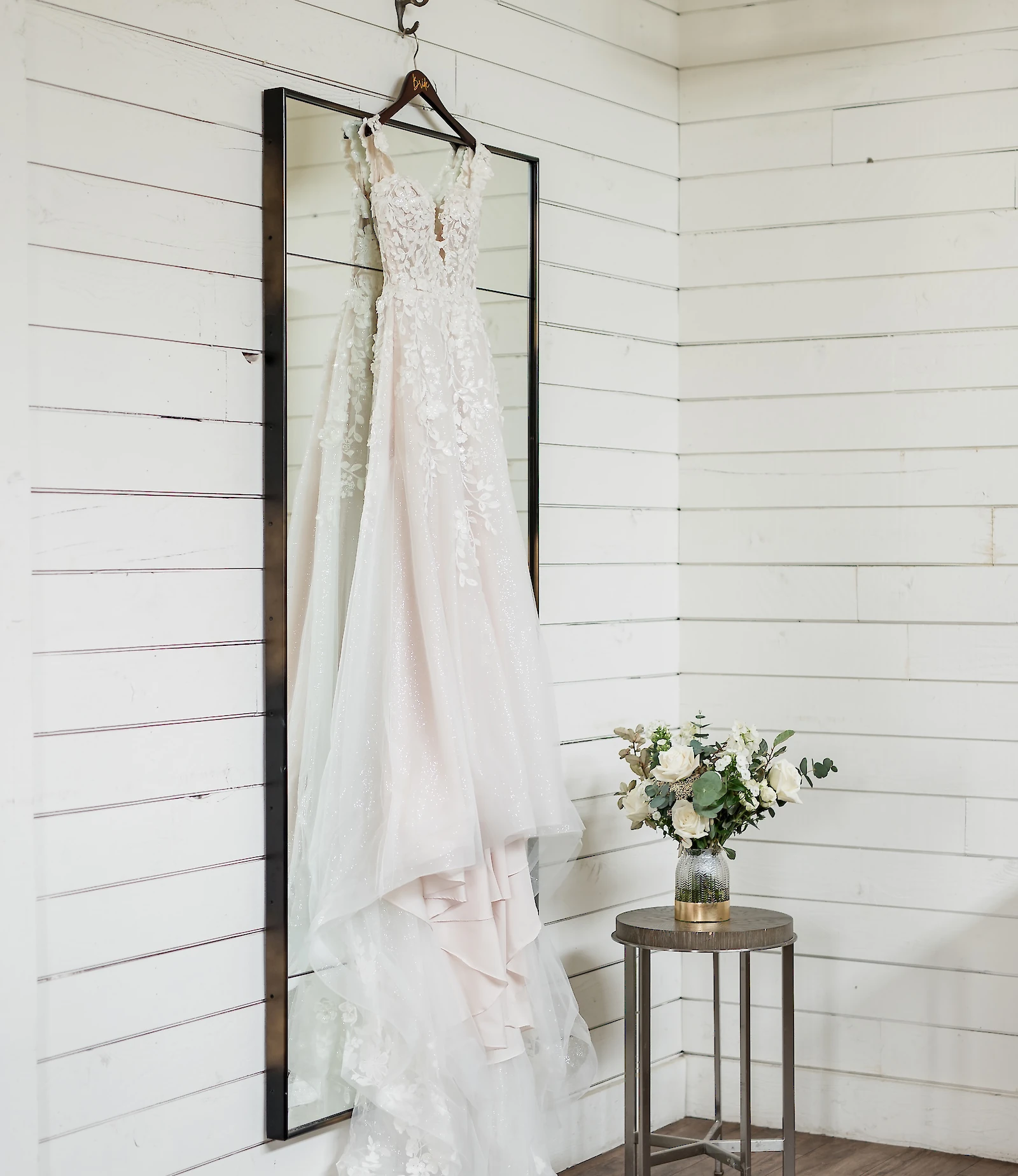 Blush-toned lace wedding dress with a deep V-neckline hanging on a mirror against a white plank wall in a bridal suite. A white and green floral bouquet rests on a small side table next to the dress.