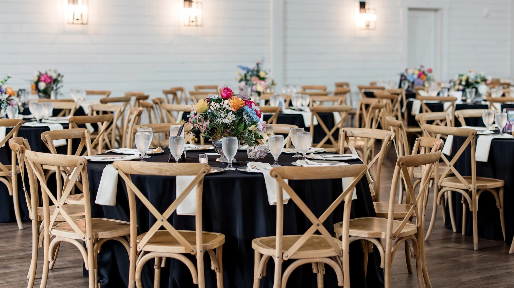 Reception hall with round tables covered in black linens, wooden cross-back chairs, and colorful floral centerpieces
