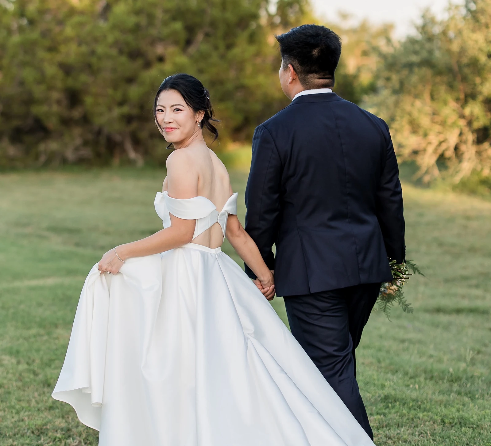 Back view of an Asian wedding couple walking away on a grassy lawn with the bride looking back over her shoulder. The bride wears a white satin off-the-shoulder gown with a long train.