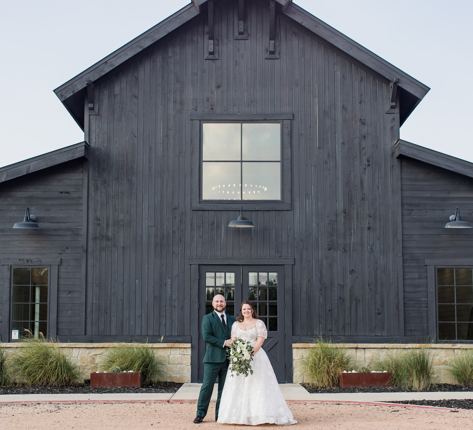 Wedding portrait of the couple standing outside a modern black barn venue. The groom wears a dark green suit, and the bride wears a cap-sleeved lace wedding dress and holds a white floral bouquet.