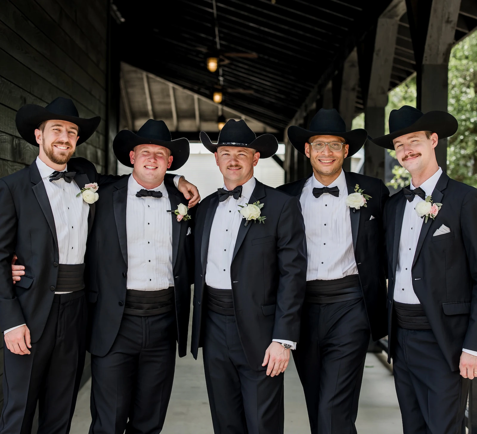 Group portrait of five groomsmen smiling and wearing classic black tuxedos with bow ties and black cowboy hats, posing under the dark wooden beams of a rustic wedding venue.