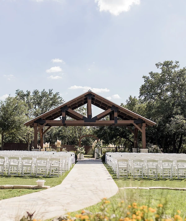 Modern rustic wedding venue entrance with a high-pitched metal roof, exposed heavy timber beams, and black metal structural plates. The porch overlooks manicured landscaping and a grassy area.