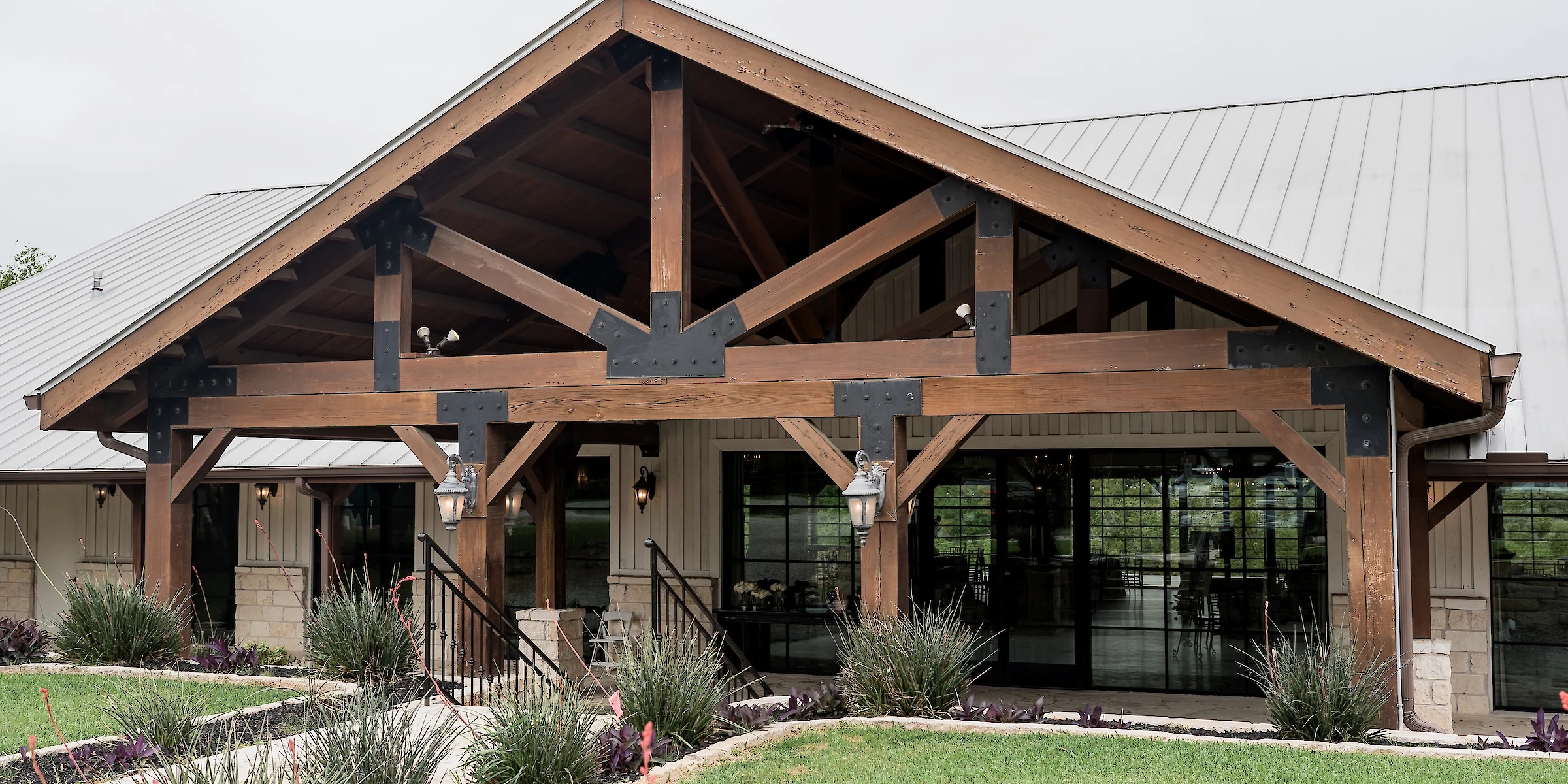 Modern rustic wedding venue entrance with a high-pitched metal roof, exposed heavy timber beams, and black metal structural plates. The porch overlooks manicured landscaping and a grassy area.