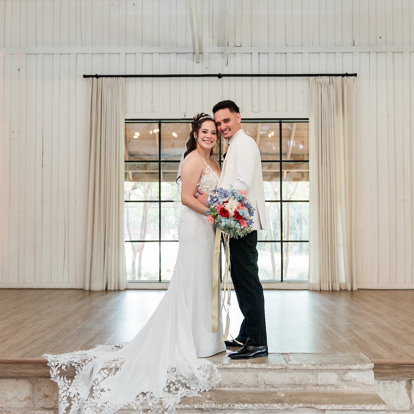 Full-length wedding portrait of the couple smiling on a stone altar in a white wood-paneled reception room. The groom wears a white tuxedo jacket and the bride holds a vibrant red and blue floral bouquet, with the lace train of her gown spread out.
