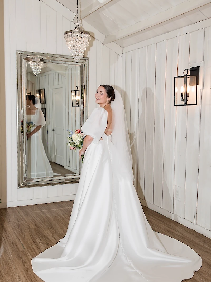 Bridal portrait of the bride looking over her shoulder in a bright white dressing suite. She wears a modern white satin wedding gown with puffy shoulder sleeves, an open back, and a full skirt, holding a colorful bouquet.
