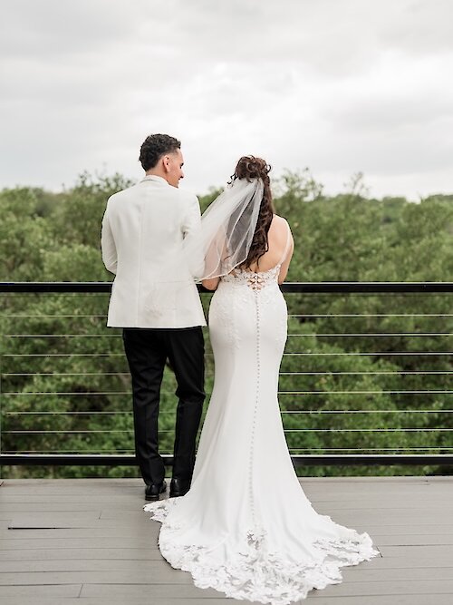 Back view of the bride and groom standing on an outdoor balcony overlooking a lush green forest. The groom wears a unique white tuxedo jacket, and the bride wears a mermaid-style dress with a button-up back and lace train.