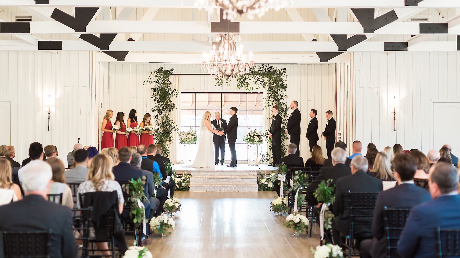 Wide view of an indoor wedding ceremony in a bright white farmhouse-style venue with exposed white wood beams and black metal accents. The altar features an arched greenery garland and the bridal party is dressed in deep red gowns.