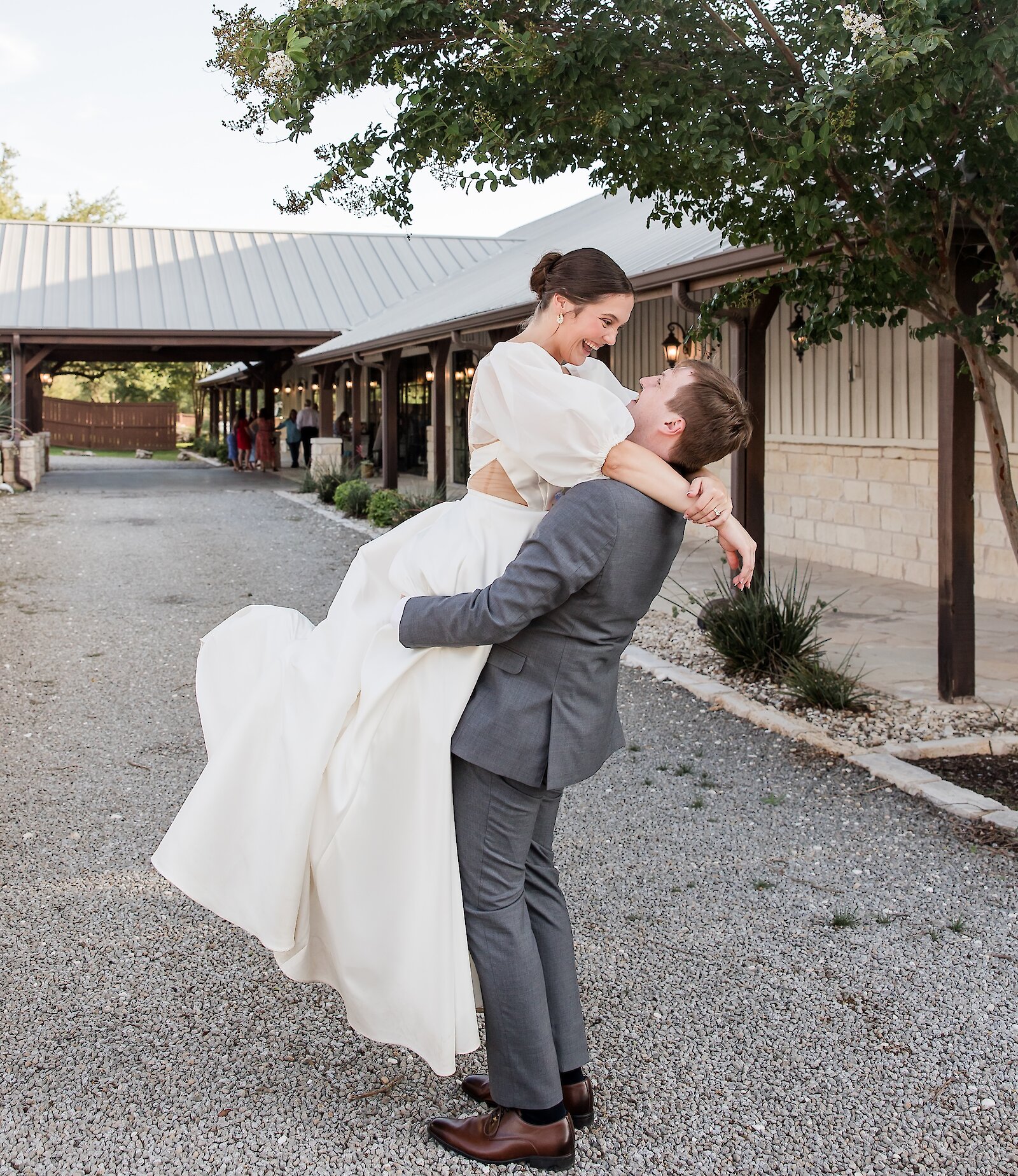 Newlywed couple celebrating outdoors on a gravel path, with the groom wearing a gray suit lifting and spinning the bride who is wearing a two-piece white satin wedding dress.