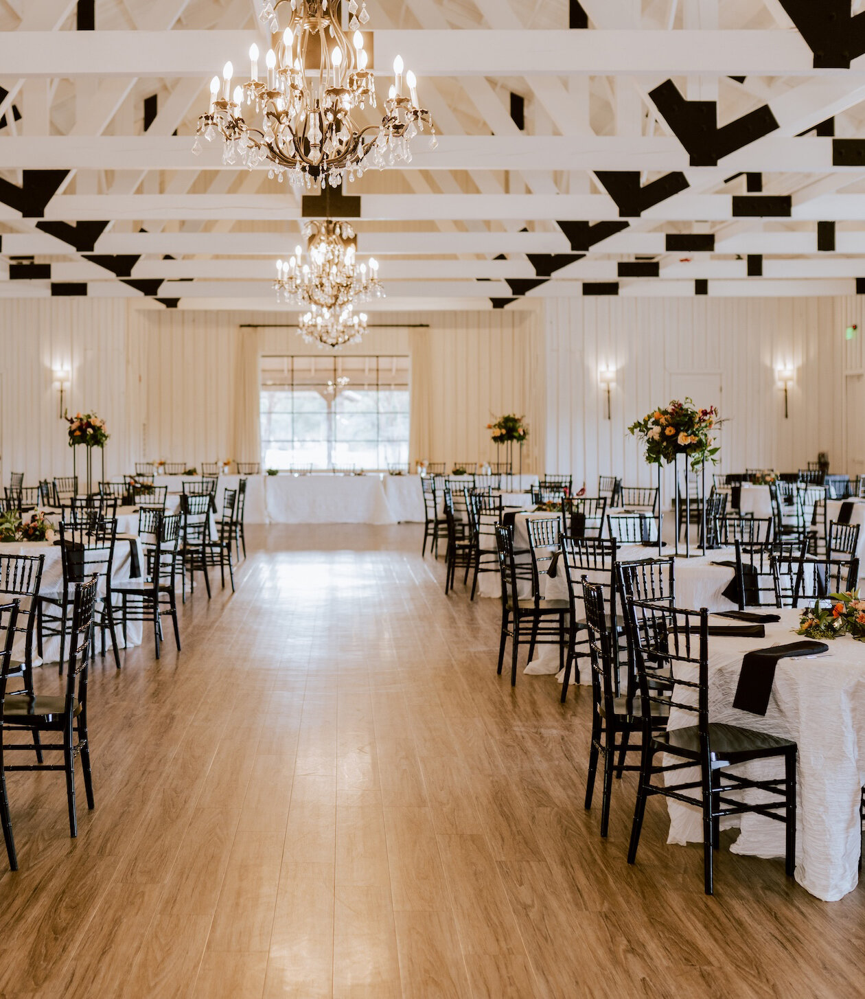 Elegant wedding reception hall with black chairs, white table linens, tall floral centerpieces, and crystal chandeliers hanging from white exposed beams