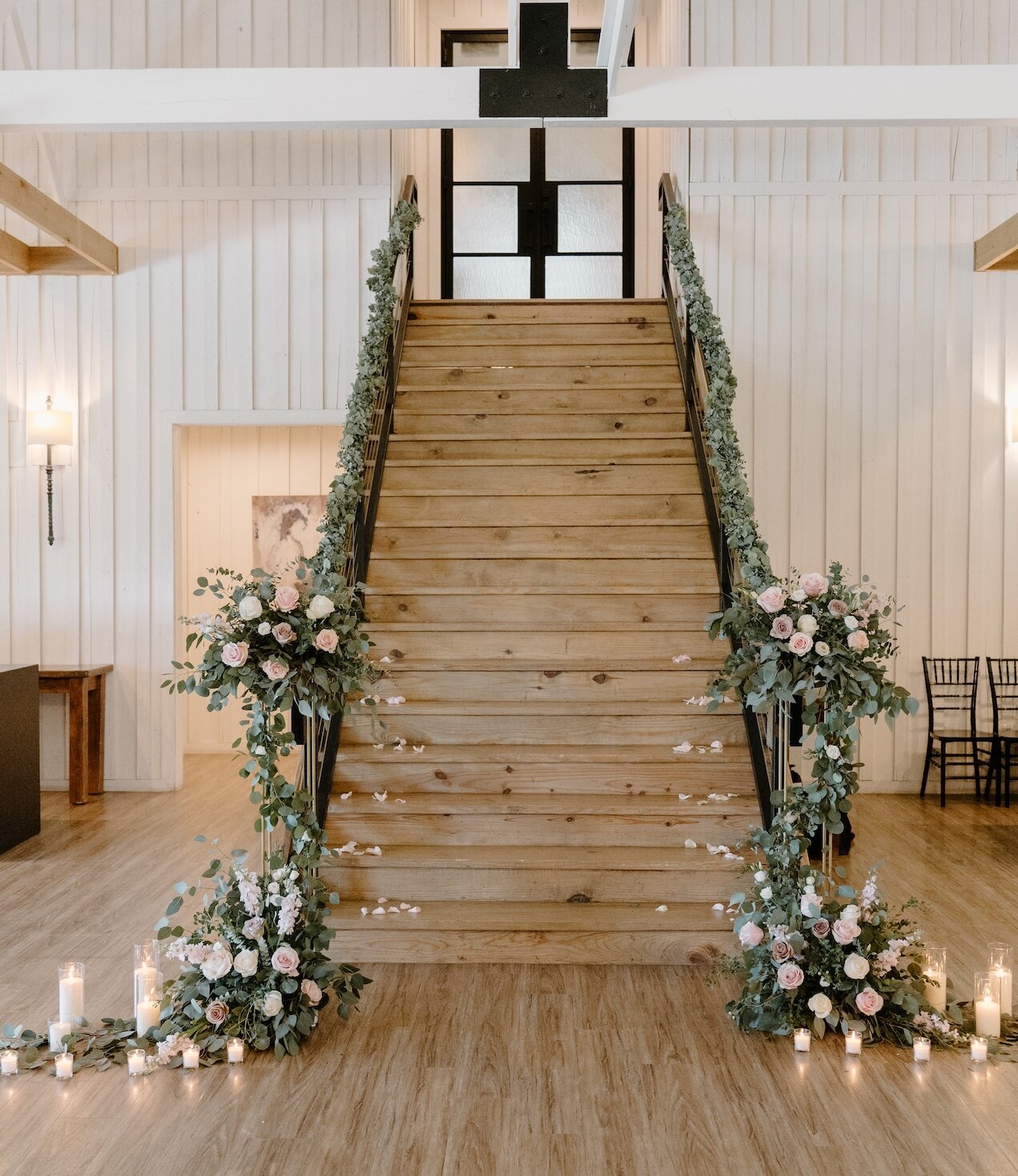 Grand wooden staircase in a white farmhouse-style wedding venue, decorated with blush pink and white floral garlands, scattered rose petals, and glowing pillar candles on the floor for a romantic ceremony accent.