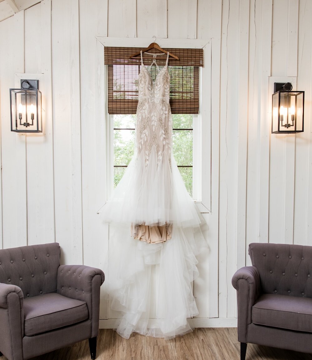 Intricate beaded lace wedding gown with a deep V-neckline and tulle skirt, hanging in a bright bridal suite window between two dark gray upholstered armchairs and rustic wall sconces.