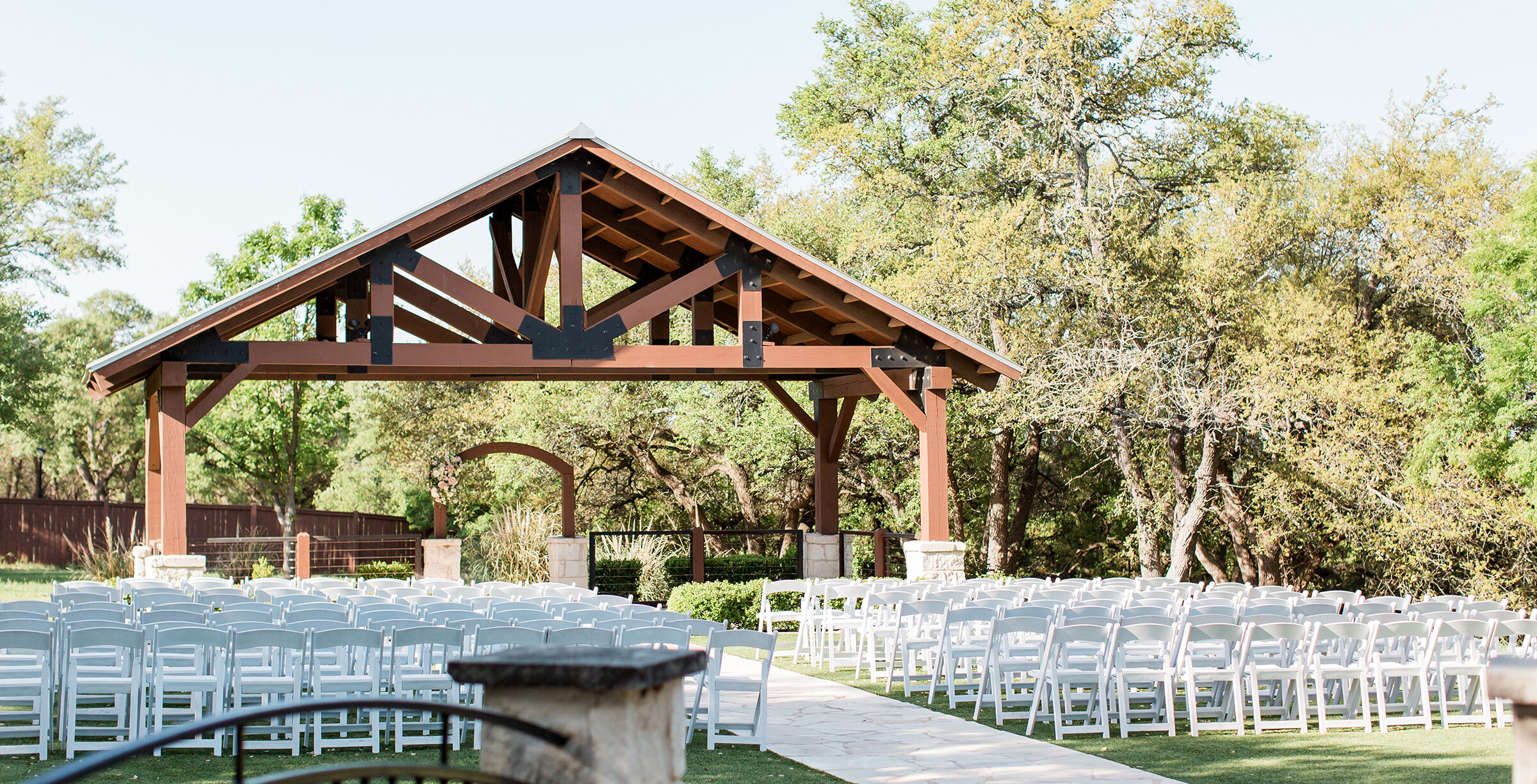 Outdoor wedding ceremony setup featuring rows of white chairs facing a wooden pavilion with exposed beams, stone columns, and surrounding trees under a clear sky.