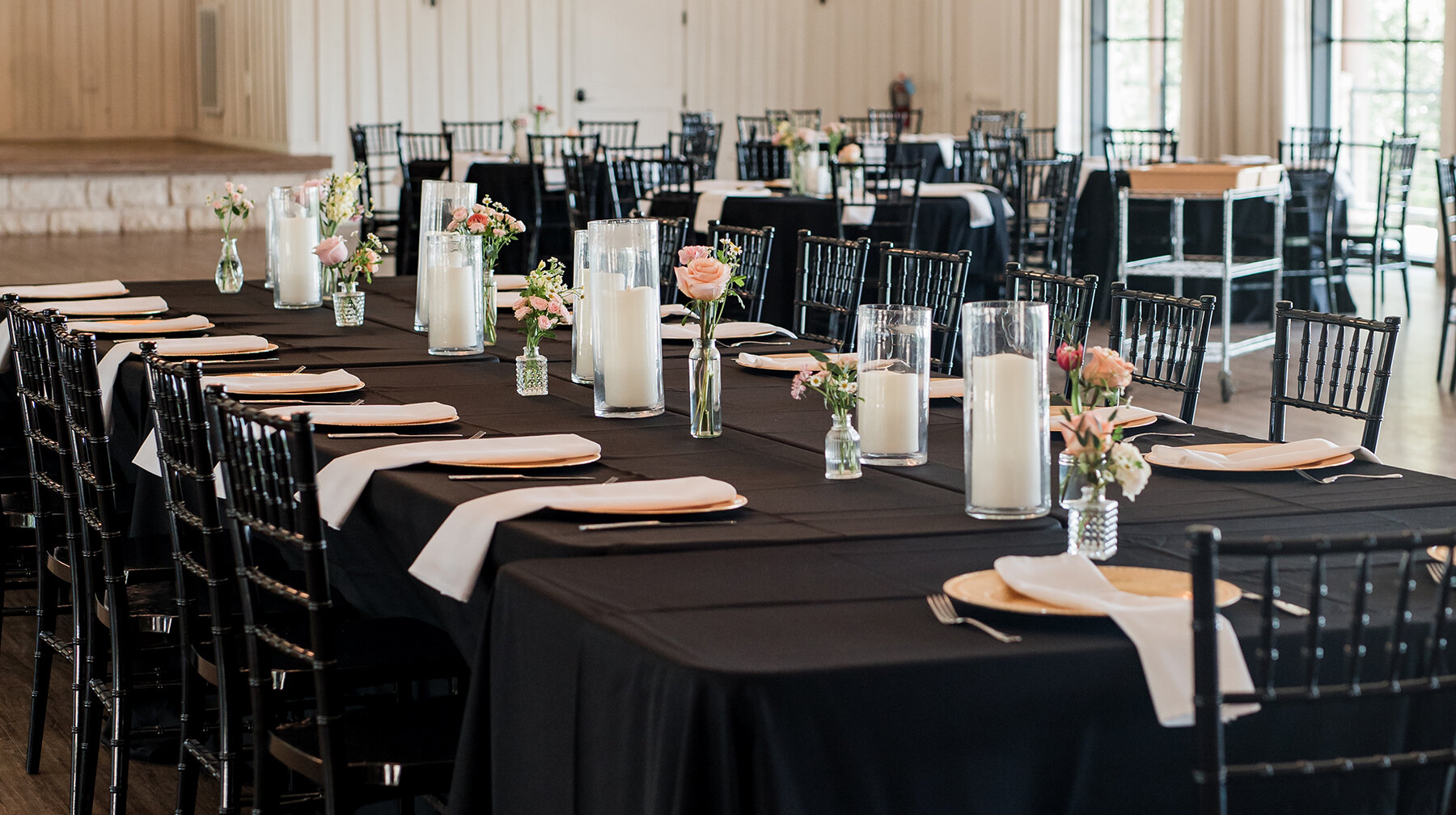 Wedding reception setup with black tablecloths, black chiavari chairs, gold chargers, white napkins, tall cylinder candles, and small floral bud vases creating a modern elegant table design