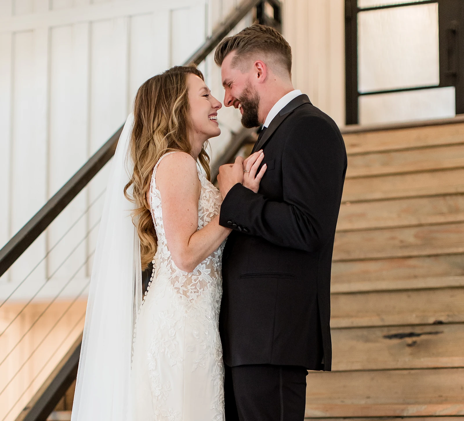 Bride and groom sharing an emotional first look moment beside a wooden staircase, smiling and holding hands in wedding attire.