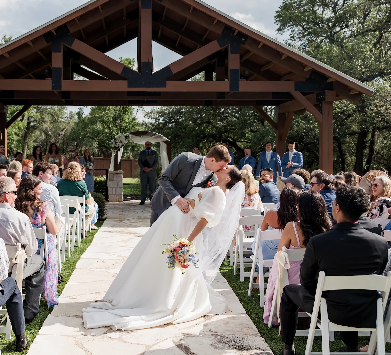 Newlywed couple sharing a dramatic kiss and dip while walking down the aisle after their outdoor ceremony under a rustic wooden pavilion. Guests are seated in white chairs on a grassy lawn.
