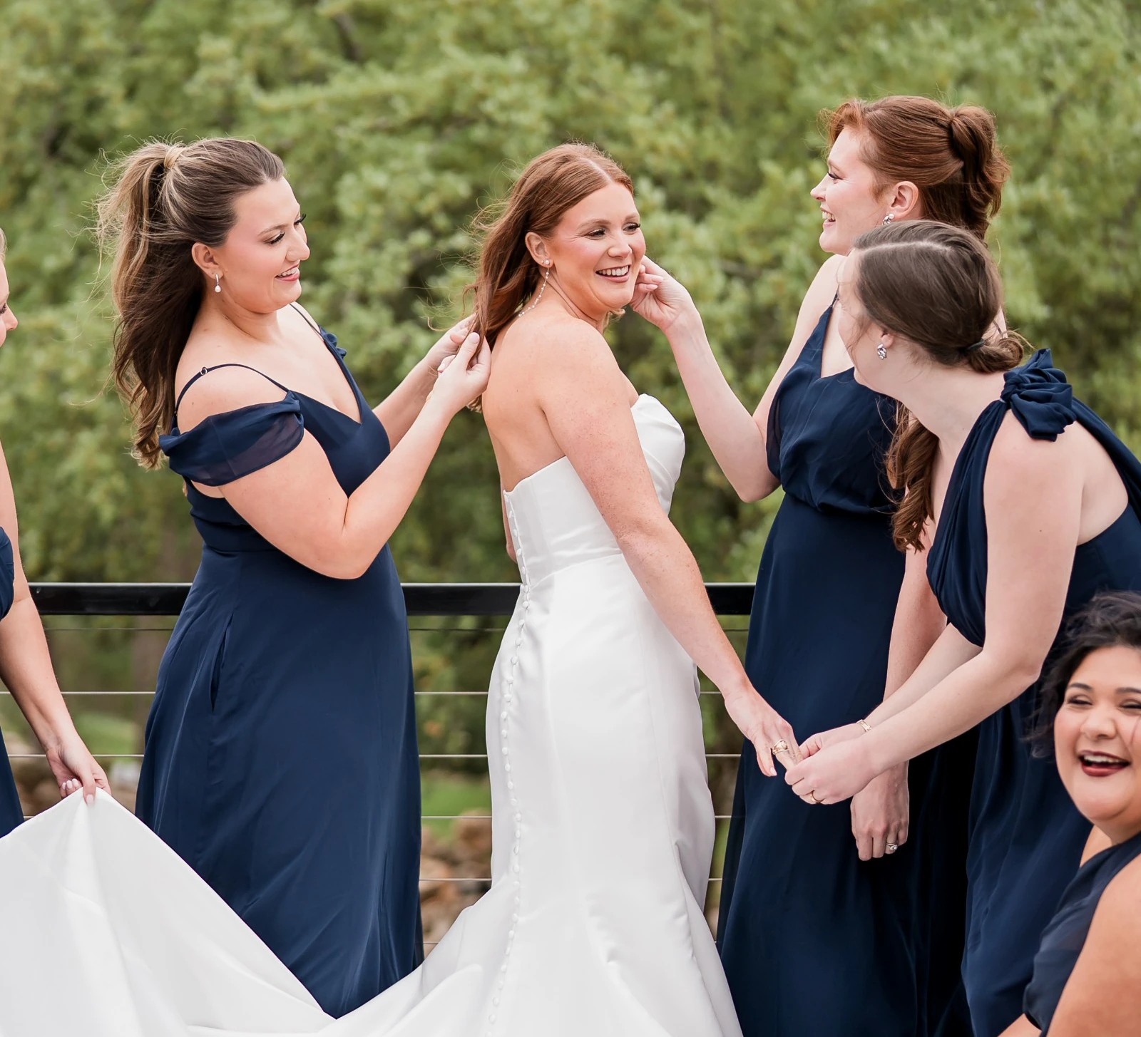 Bride and bridesmaids posing on an outdoor deck as the bridesmaids adjust the strapless white satin gown and spread out the skirt. The bridesmaids are wearing matching navy blue dresses with varying necklines.