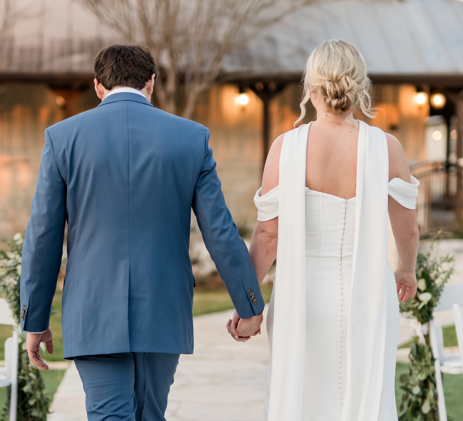 Back view of a newlywed couple walking hand-in-hand down a flagstone aisle lined with white chairs and greenery garlands after their ceremony. The groom wears a blue suit, and the bride wears a unique white gown with shoulder capes and a buttoned back.