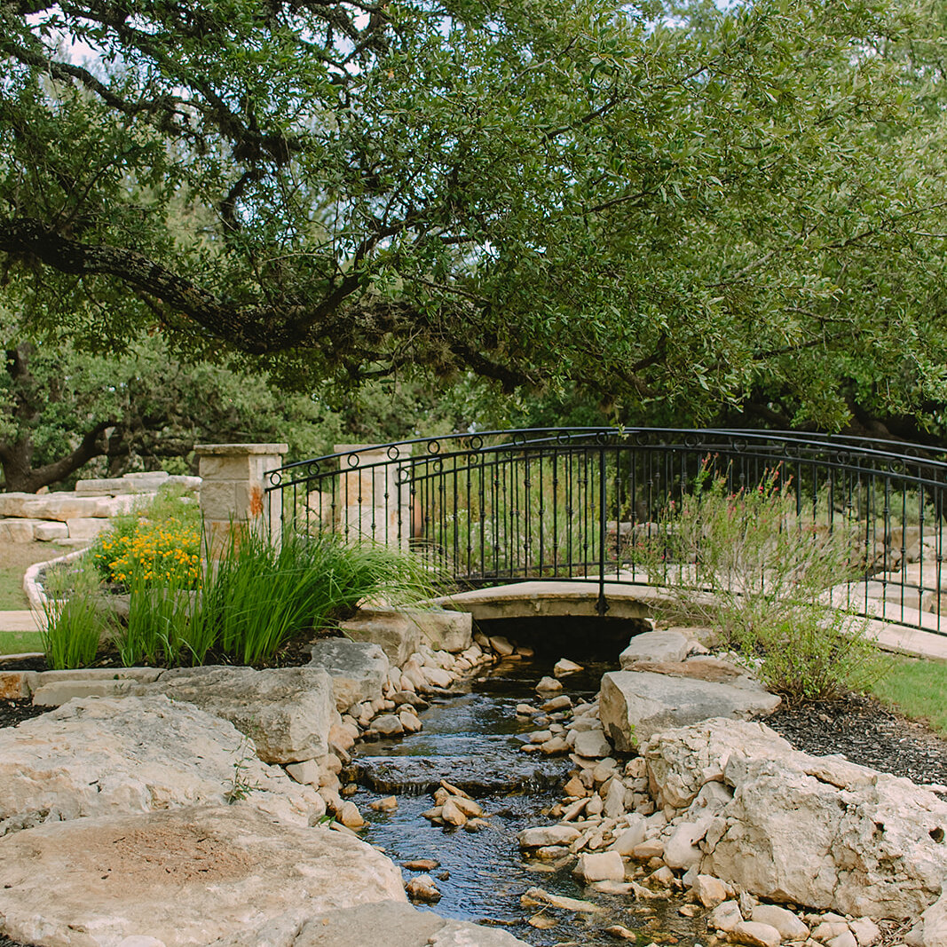 Scenic outdoor garden pathway featuring a small black iron arch bridge crossing a flowing rock creek, surrounded by large oak trees and natural landscaping.