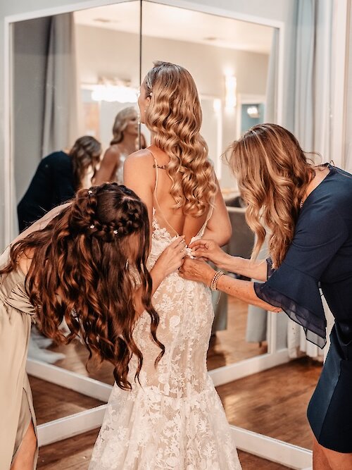 Intimate moment of the bride getting buttoned into her lace wedding dress by her mother and maid of honor in the bridal suite. The bride has long blonde curled hair and a low, buttoned back on her gown.