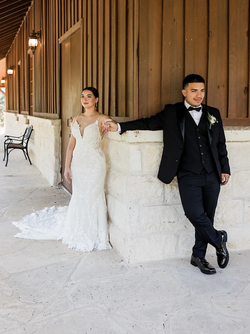 Stylish wedding portrait of an elegant couple posing on a stone porch of a rustic venue with vertical wood siding. The groom wears a black tuxedo and the bride wears a plunging lace mermaid gown.