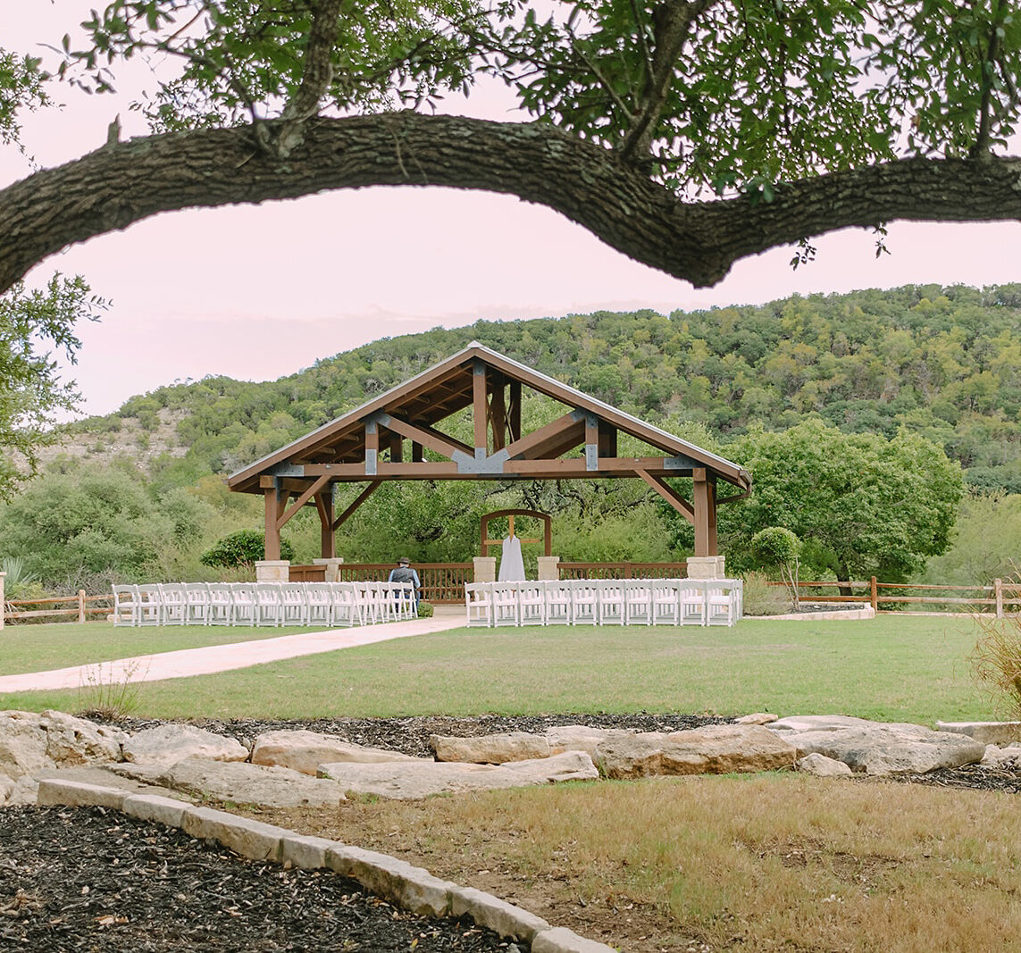 Outdoor wedding ceremony setup featuring a wooden pavilion, white chairs, and a scenic Hill Country backdrop framed by large tree branches