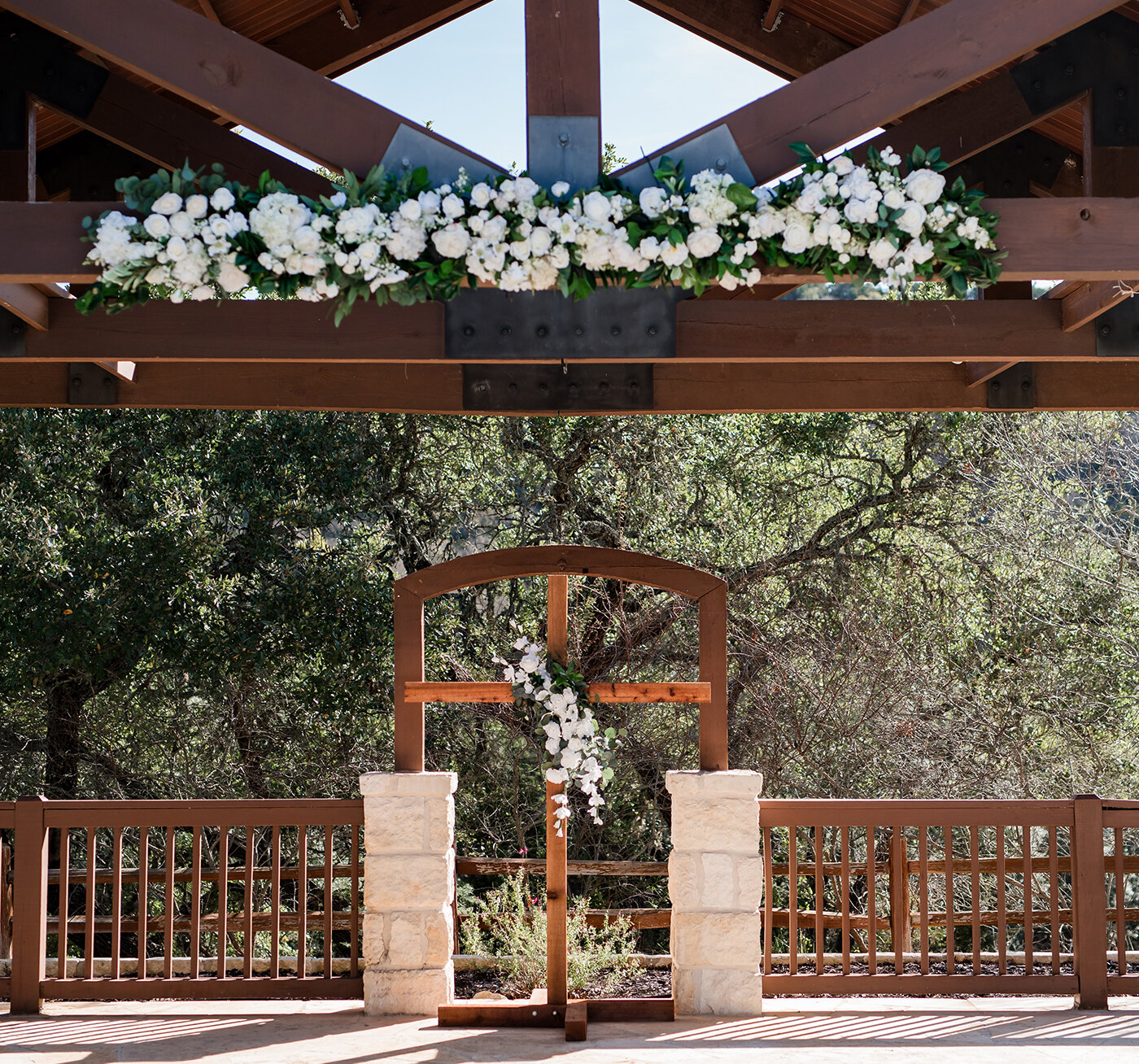 Wooden cross wedding ceremony altar decorated with white flowers beneath a rustic pavilion surrounded by trees