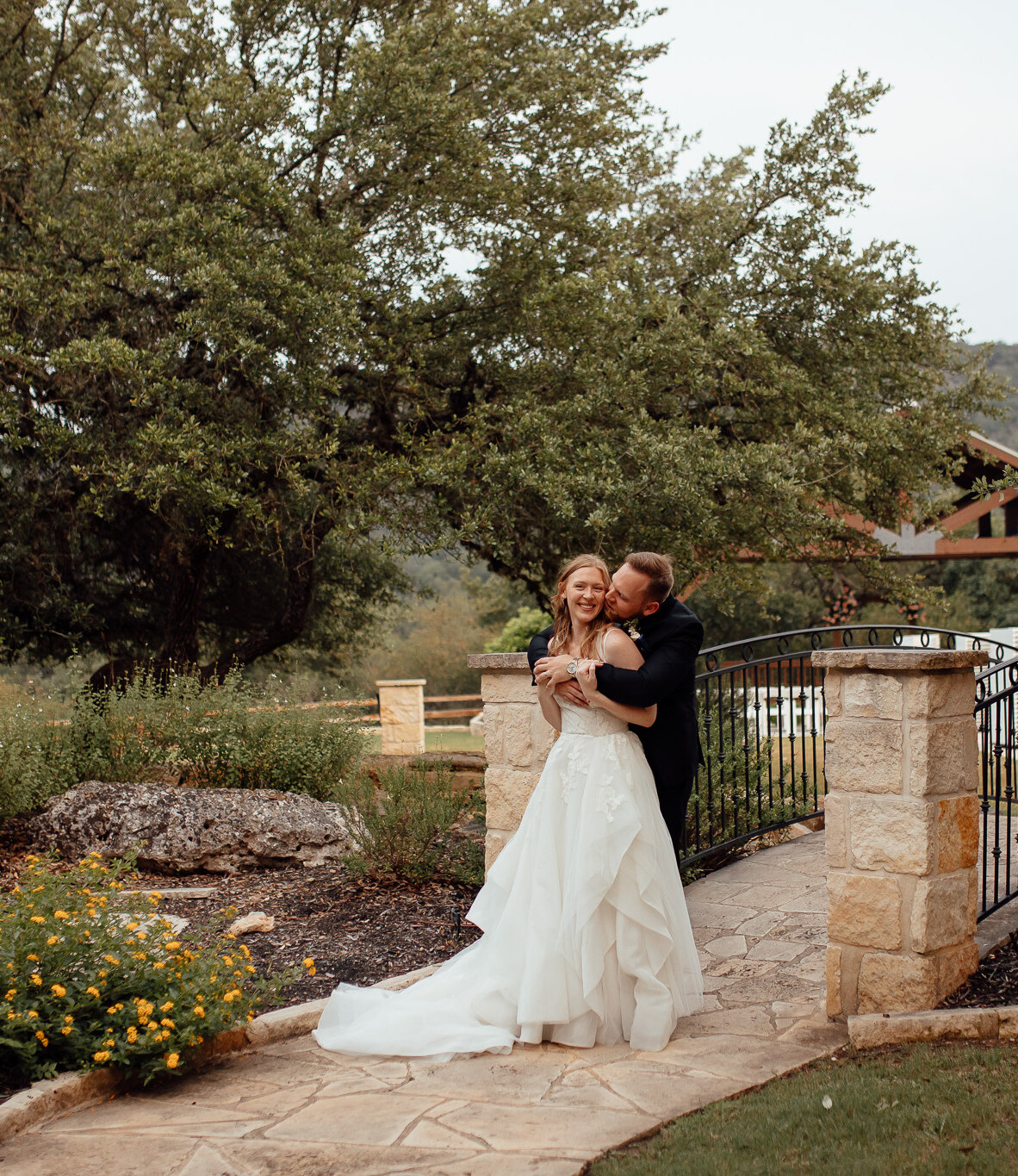 Bride and groom embracing and laughing on a stone bridge surrounded by lush gardens and oak trees at a Texas Hill Country wedding venue