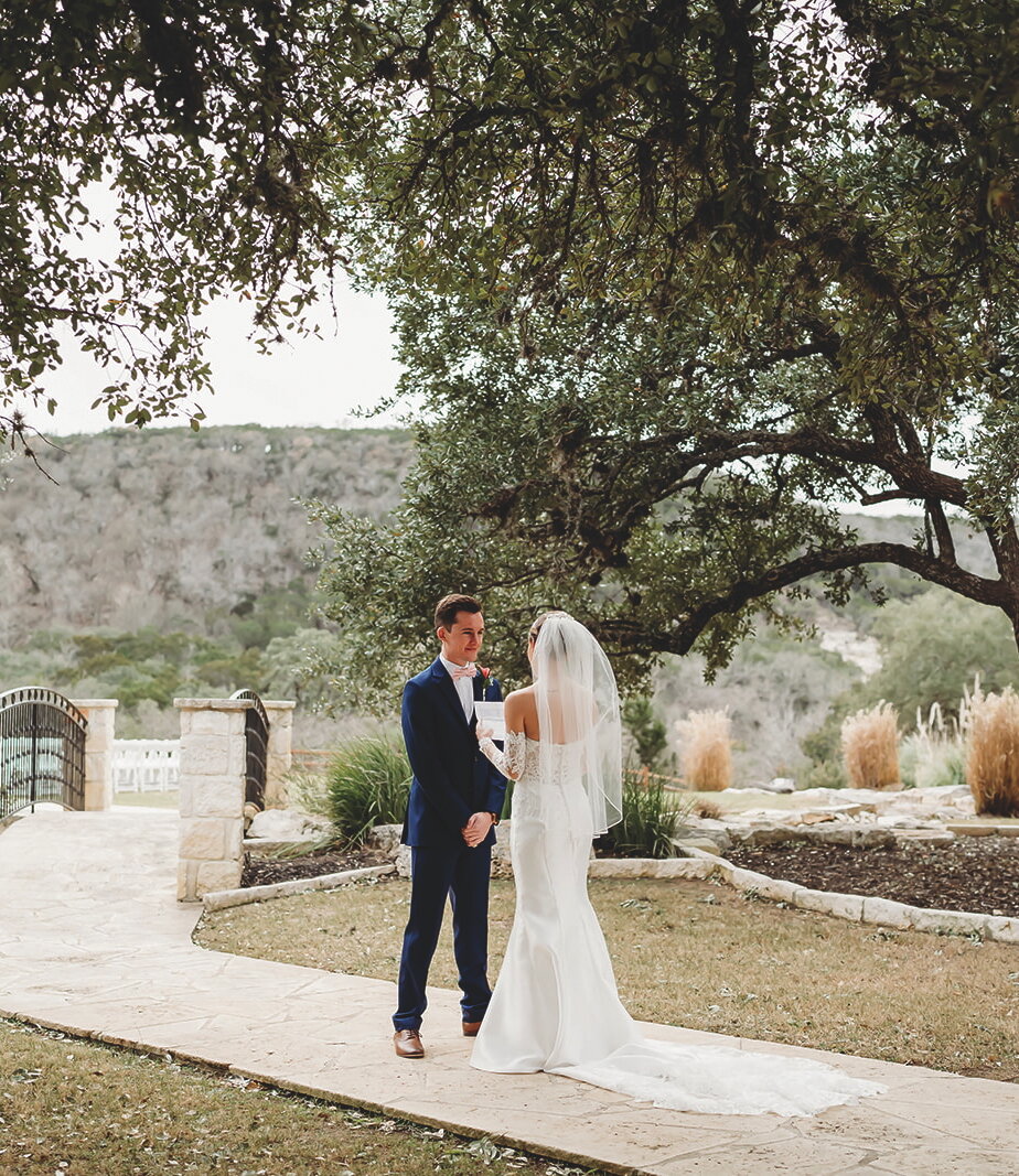 Wedding portrait of the couple facing each other on a flagstone pathway beneath large oak trees. The groom wears a navy suit and the bride wears a long-sleeved lace gown with a dramatic train, set against a hill country backdrop.