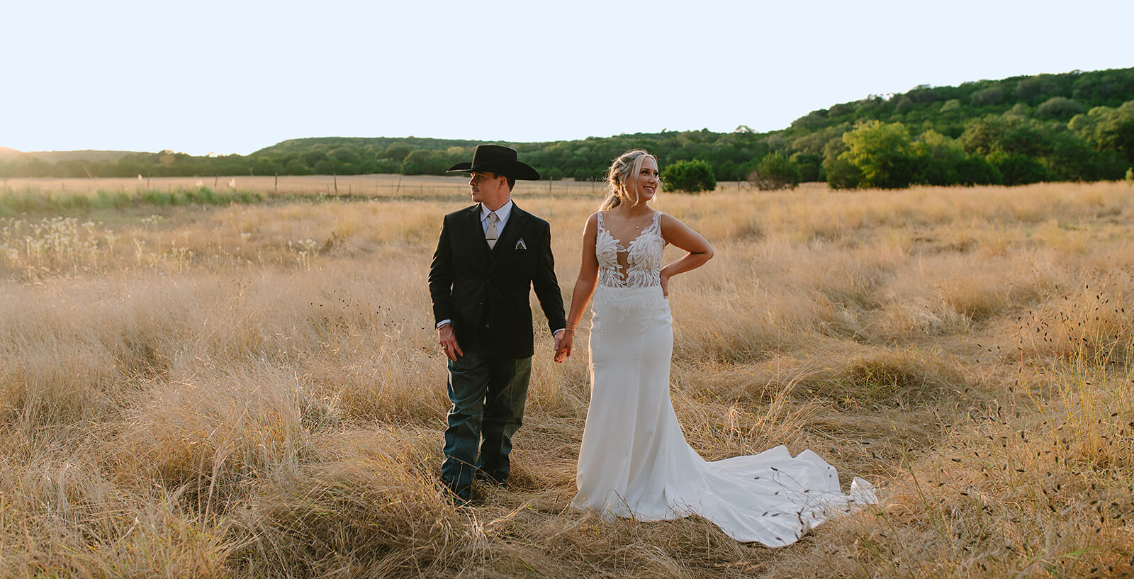 Golden hour wedding portrait of the couple walking hand-in-hand through a tall, dry grass field. The groom wears a black suit, denim jeans, and a black cowboy hat, and the bride wears a lace mermaid-style gown.