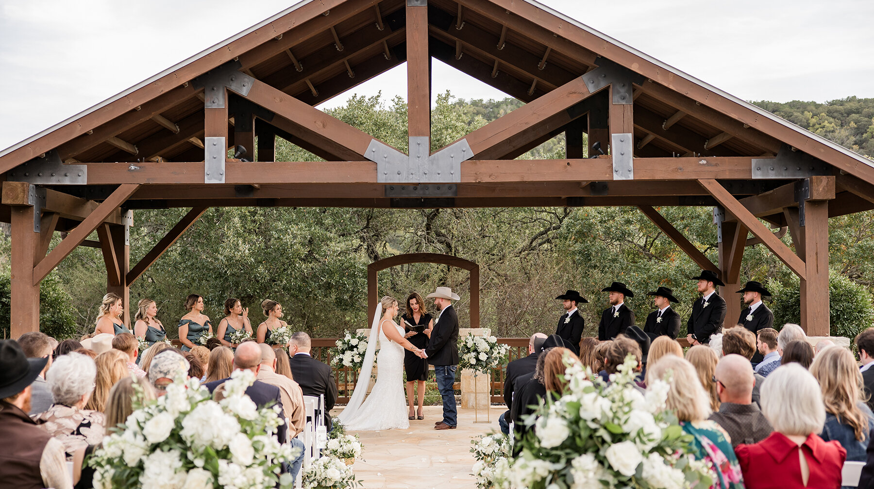 Bride and groom exchanging vows under a wooden pavilion during an outdoor Texas wedding ceremony with bridesmaids, groomsmen in cowboy hats, and guests seated among white floral arrangements