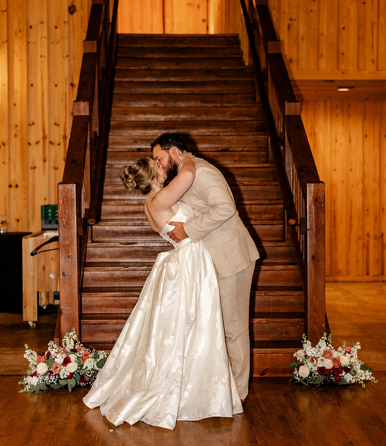 Romantic portrait of the bride and groom kissing and dipping on a wooden dance floor in front of a grand wooden double staircase. The rustic barn venue features wood-plank walls and low floral arrangements with red and white roses.