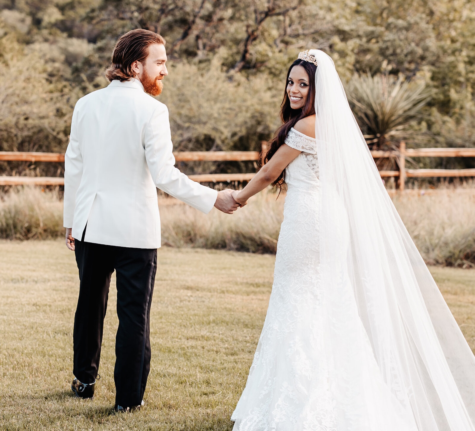 Romantic wedding portrait of the bride and groom walking away across a grassy field with the bride looking back and smiling. The groom wears a white tuxedo jacket and the bride wears a lace off-the-shoulder gown with an elaborate cathedral veil and tiara.