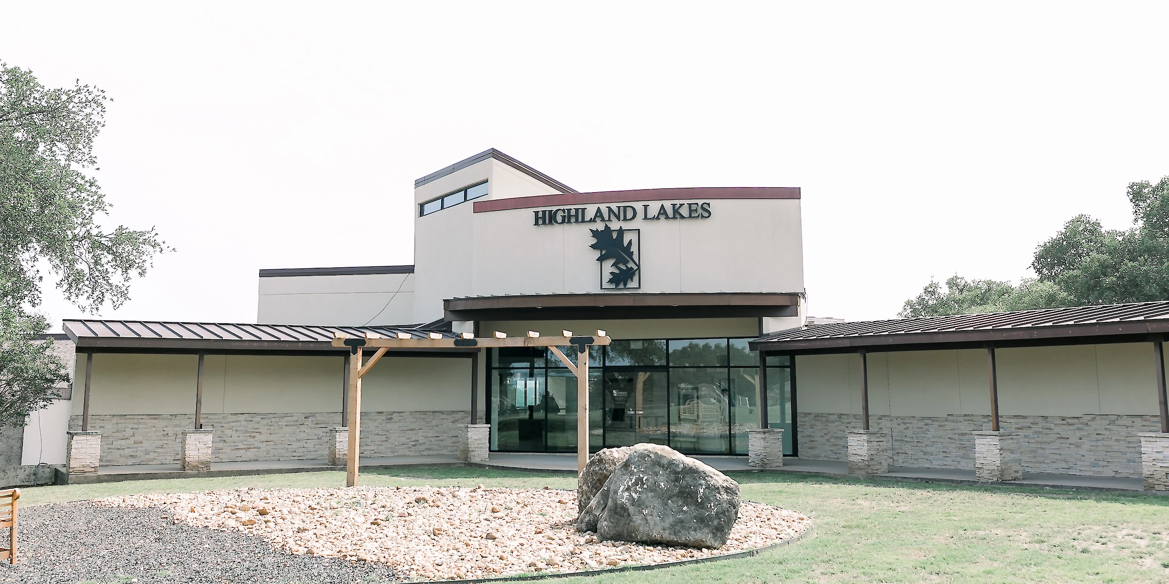 Exterior of the Highland Lakes event venue featuring a modern stucco building entrance with large glass windows and an extended covered porch area, set on a lawn with rock landscaping.