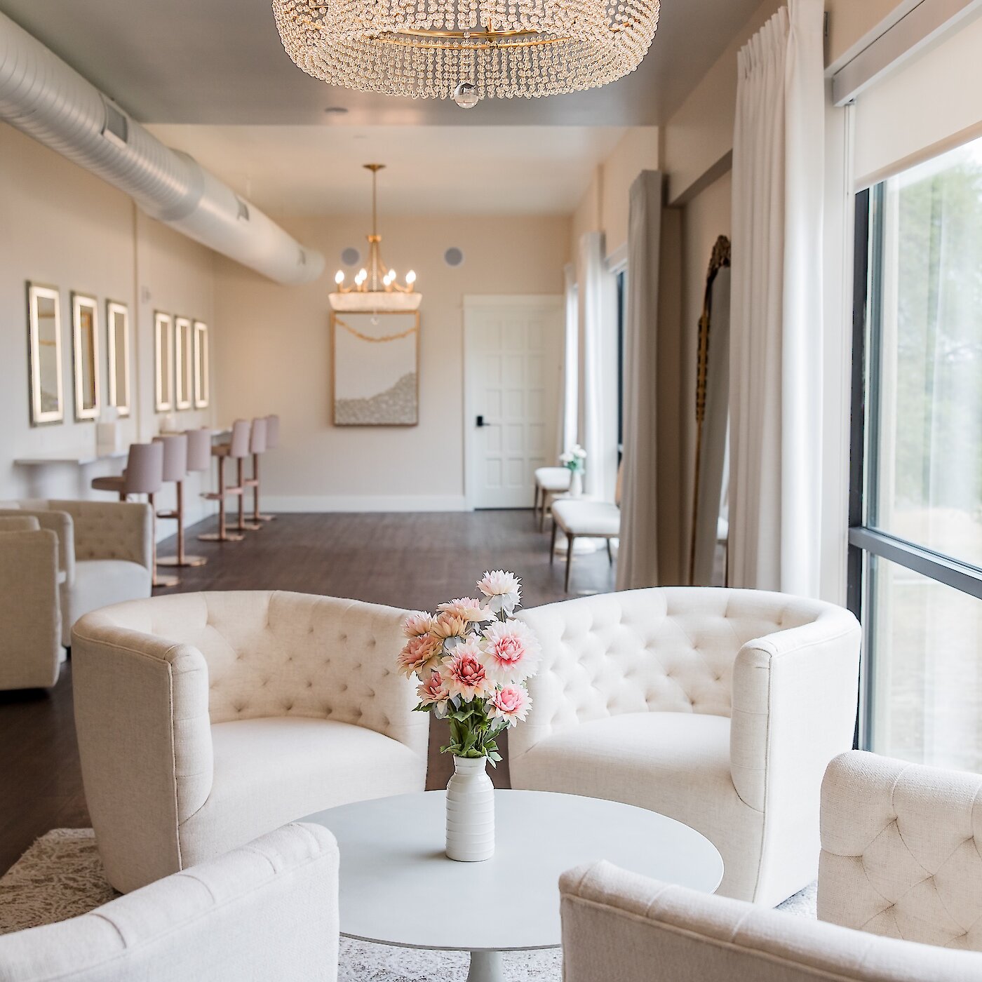 Bright and elegant bridal suite or lounge with tufted white armchairs, dark wood flooring, and a large window. The background features a vanity area with pink bar stools and a large crystal chandelier overhead.