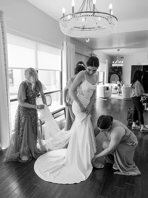 Black and white photo of the bride getting dressed in a bright bridal suite with a chandelier. The mother of the bride arranges the veil while a bridesmaid helps the bride with her shoes and the fit of her sleek satin wedding gown.