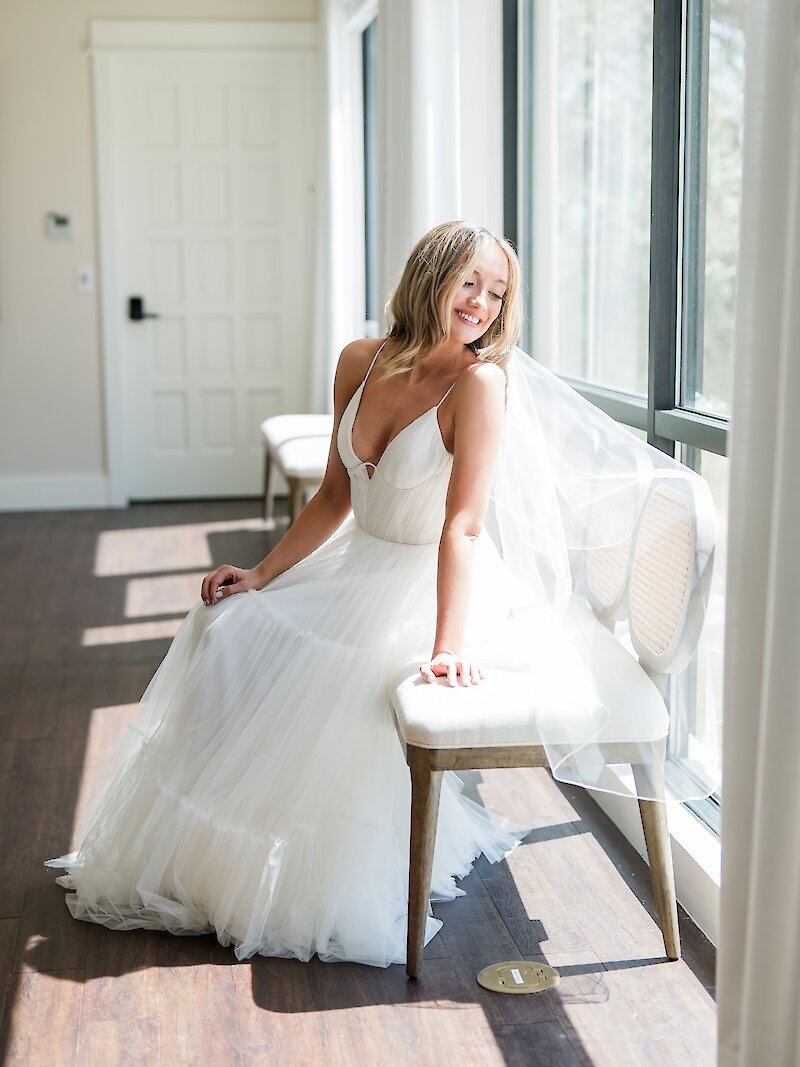 Bright bridal portrait of the bride smiling while seated in a sunlit window. She wears a white gown with a spaghetti-strap V-neckline and a tiered tulle skirt, with her veil softly blowing in the light.