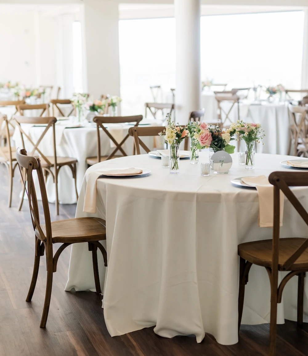 Bright wedding reception table setup with white round tablecloths, light wood cross-back chairs, and simple centerpieces featuring blush and white roses in clear bud vases. The venue has large windows and dark wood flooring.
