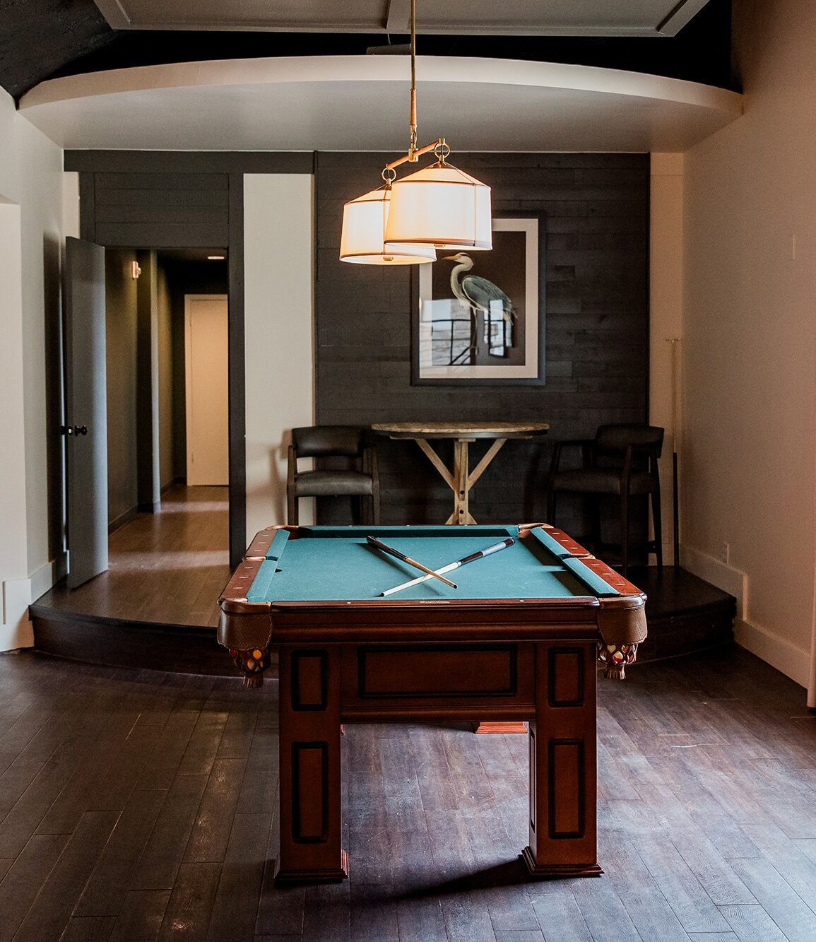 Groom’s lounge with a wooden pool table, cue sticks, dark wood floors, and modern seating area under warm lighting