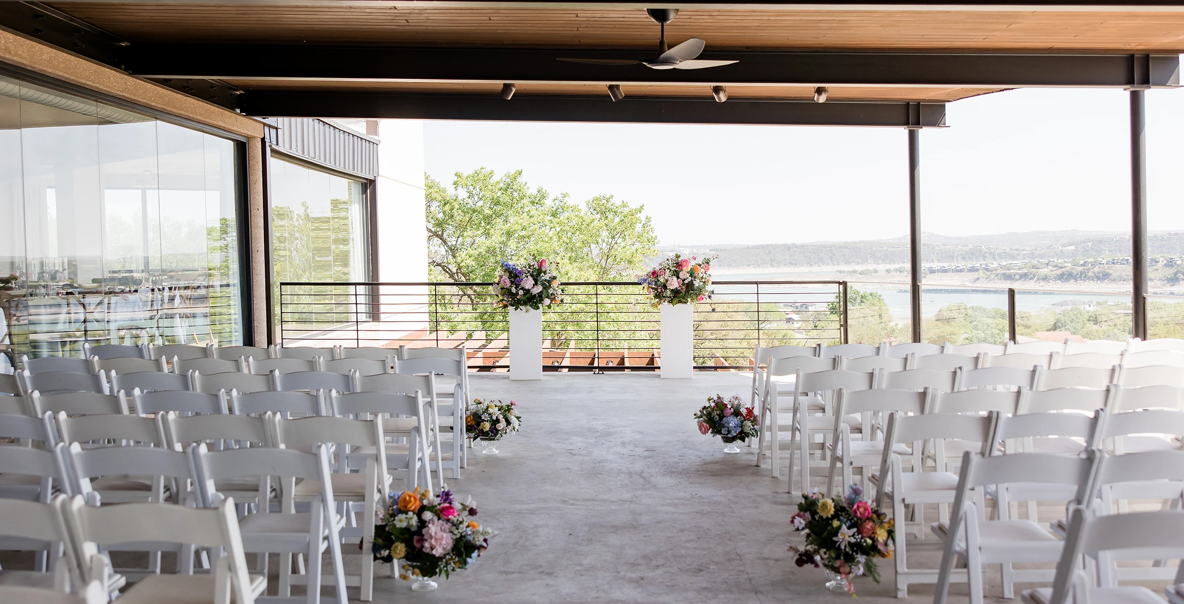Outdoor wedding ceremony setup with rows of white chairs, colorful floral arrangements, and a scenic lake view in the background