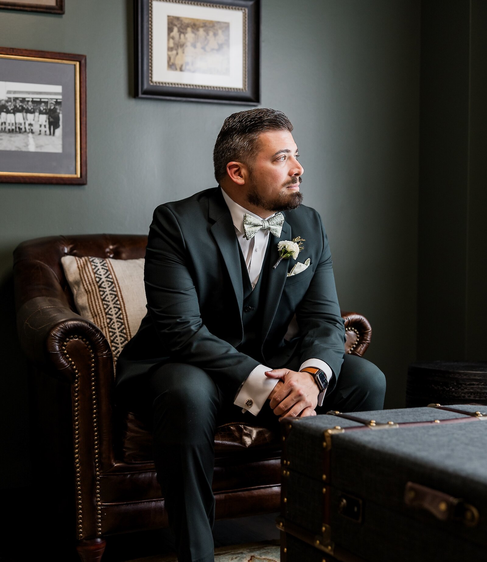 Elegant portrait of the groom seated in a leather armchair in a richly decorated room with dark green walls. He wears a fitted dark green three-piece suit with a patterned bowtie and brown leather shoes, looking thoughtfully off-camera.