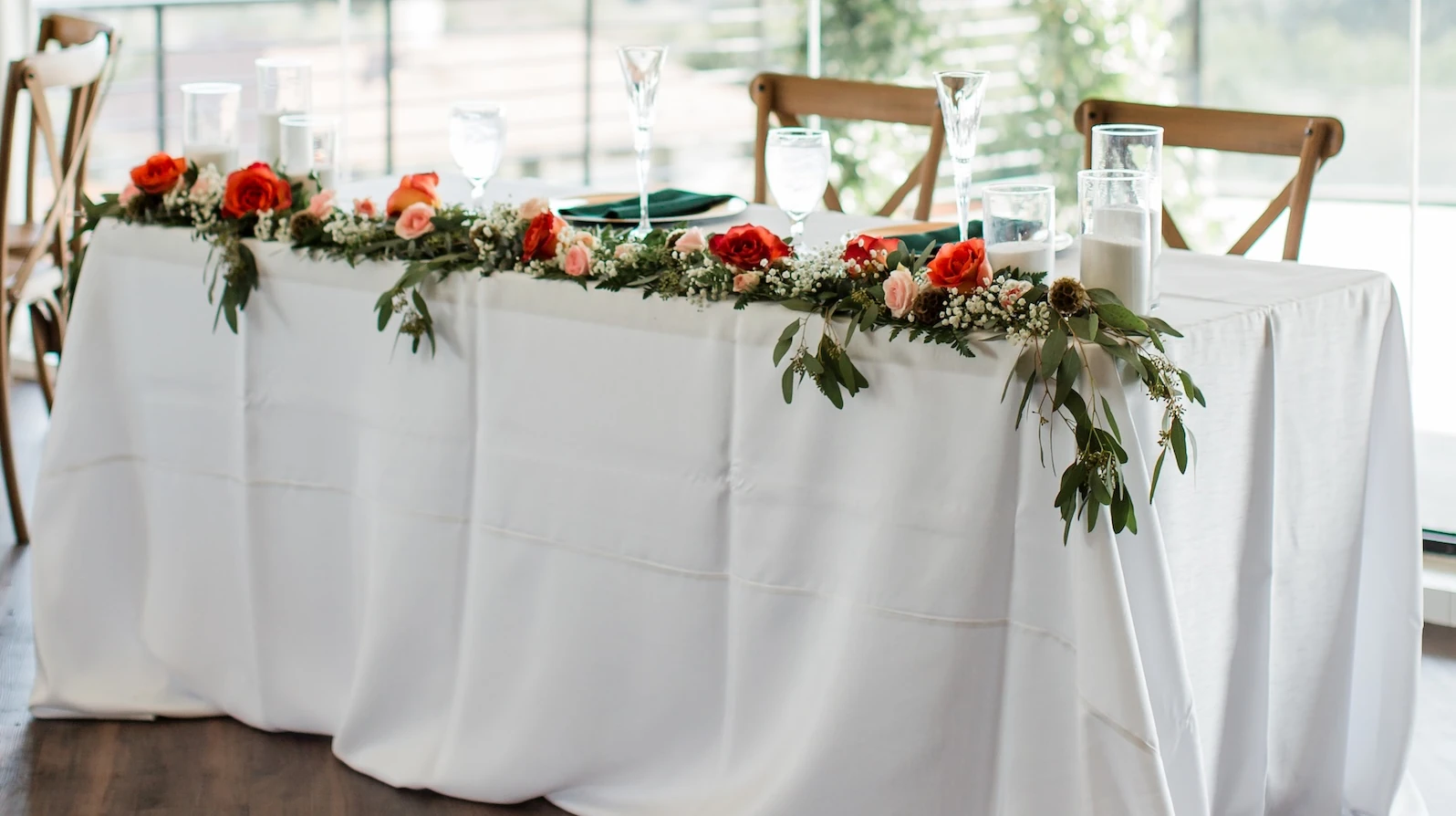 Elegant wedding sweetheart table draped in white linen, decorated with a floral garland of red and pink roses, greenery, and cylinder candles