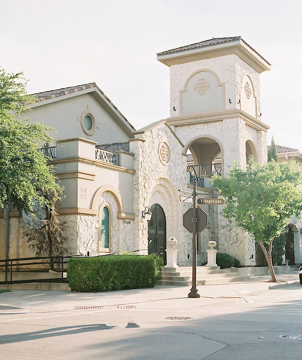 Close-up exterior of a Mediterranean-style wedding chapel featuring a prominent tower with a second-level arched balcony and rough-cut light stone facade, framed by cypress trees and wrought iron details.