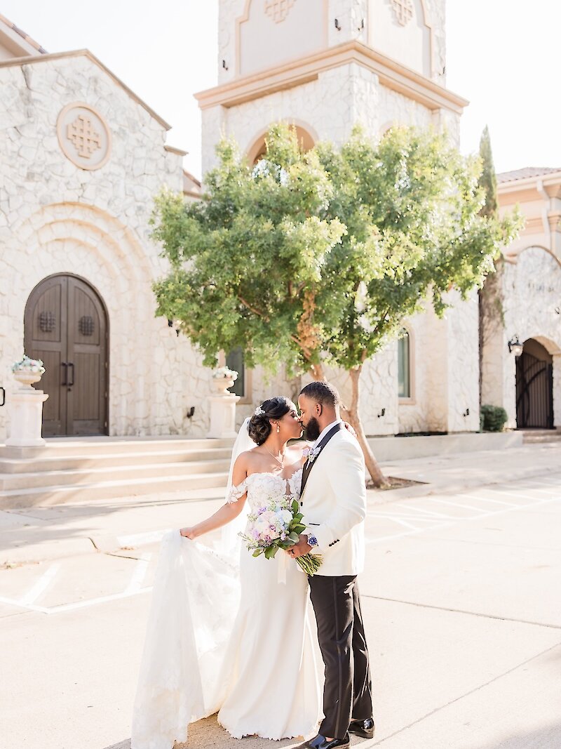 Sunny wedding portrait of the bride and groom nose-to-nose outside a Mediterranean stone chapel with a bell tower. The groom wears a white dinner jacket and the bride wears an off-the-shoulder gown, holding a purple and white bouquet.