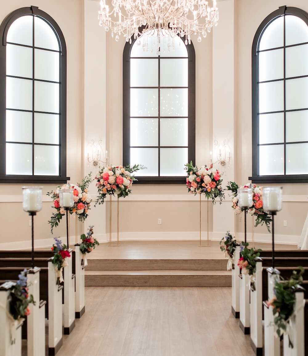 Elegant wedding chapel interior featuring a vaulted ceiling, a large crystal chandelier, and tall black-framed arched windows. The altar is decorated with vibrant pink and coral floral arrangements on gold stands, and the aisle is lined with candles and smaller coordinating flowers.