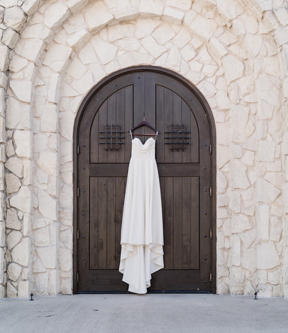 Simple, elegant strapless white wedding dress with a dramatic train, hanging against dark wooden arched doors. The doors are set into a rough-cut white stone exterior wall with an arched stone frame.