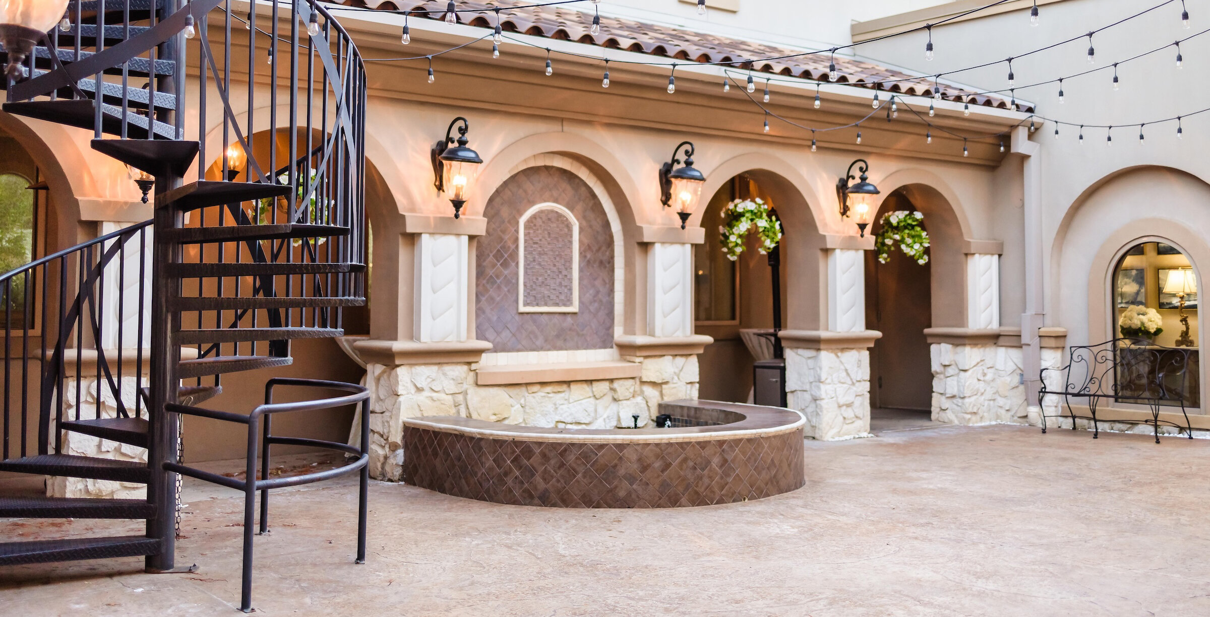 Mediterranean-style courtyard featuring arched columns, a black spiral staircase, string lights, and a small fountain area