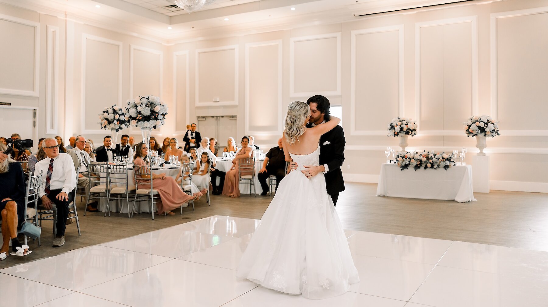Elegant wedding first dance photo in a ballroom with white paneled walls and a white dance floor. The groom embraces the bride, who wears a full ballgown, under a large circular crystal chandelier, surrounded by guests and high white floral centerpieces.