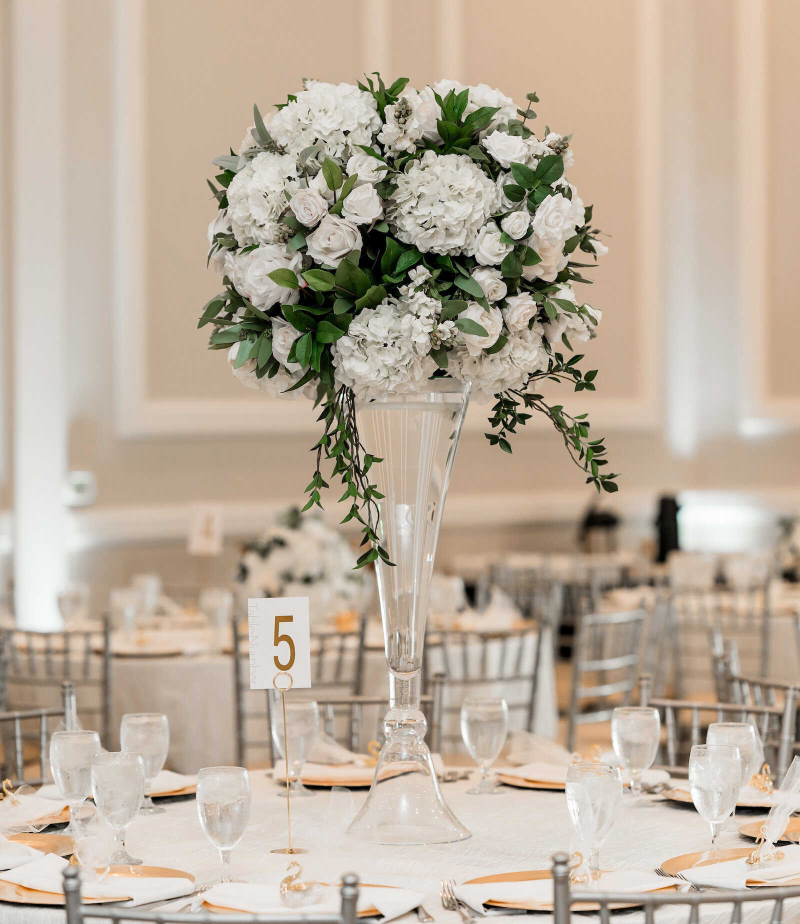 Tall glass vase with an elegant white floral centerpiece featuring roses and hydrangeas on a wedding reception table with silver chairs and table number five