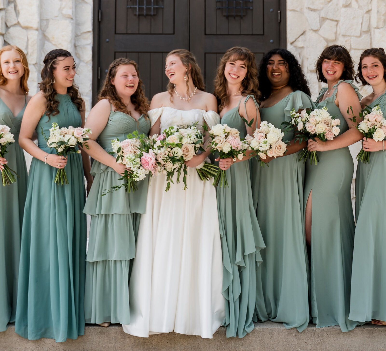 Joyful bridal party portrait of the bride and seven bridesmaids standing on stone steps outside a rough-cut stone chapel with dark arched wooden doors. The bridesmaids wear matching dusty sage green chiffon dresses and hold blush and white floral bouquets.