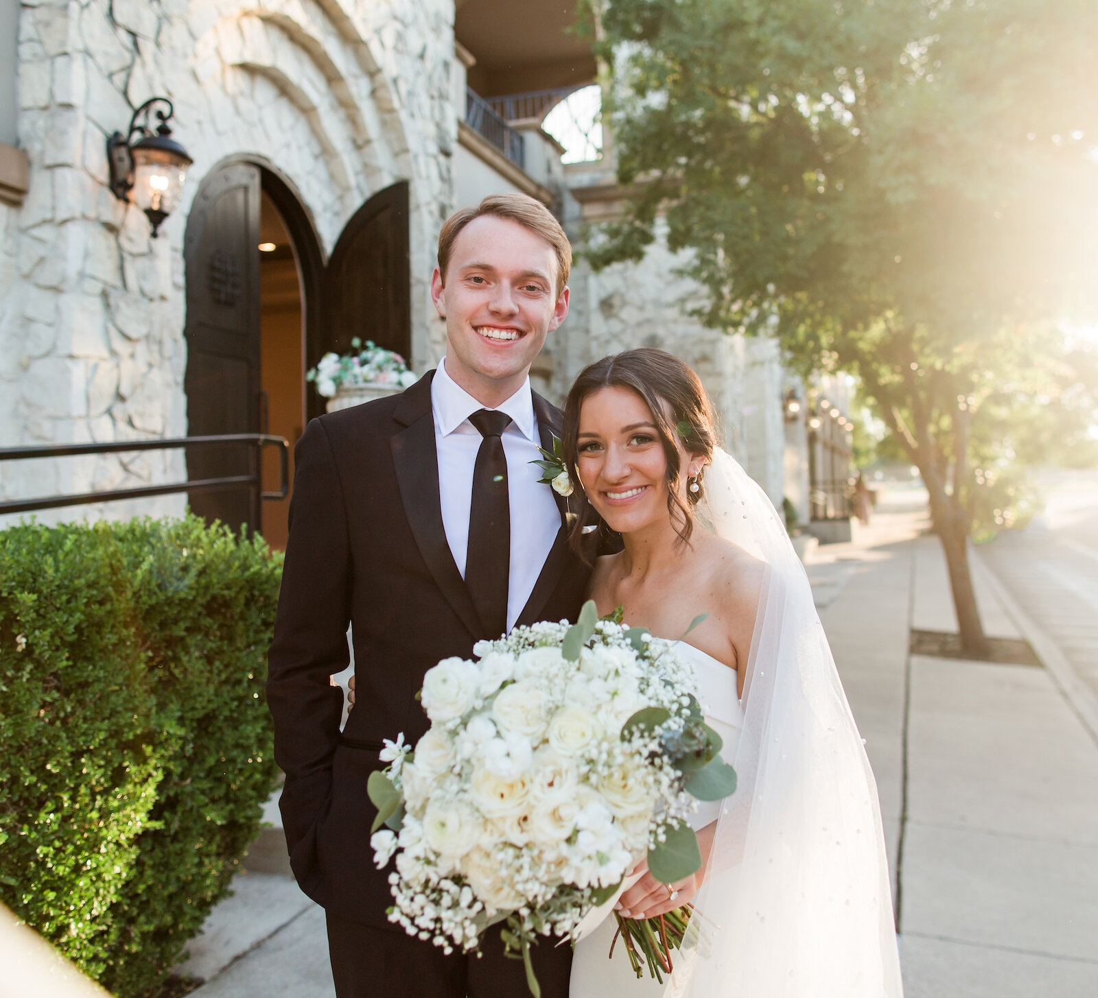 Sunlit portrait of a smiling couple standing outside a stone Mediterranean-style chapel. The groom wears a black tuxedo and the bride wears a strapless white gown, holding a large white rose and greenery bouquet.